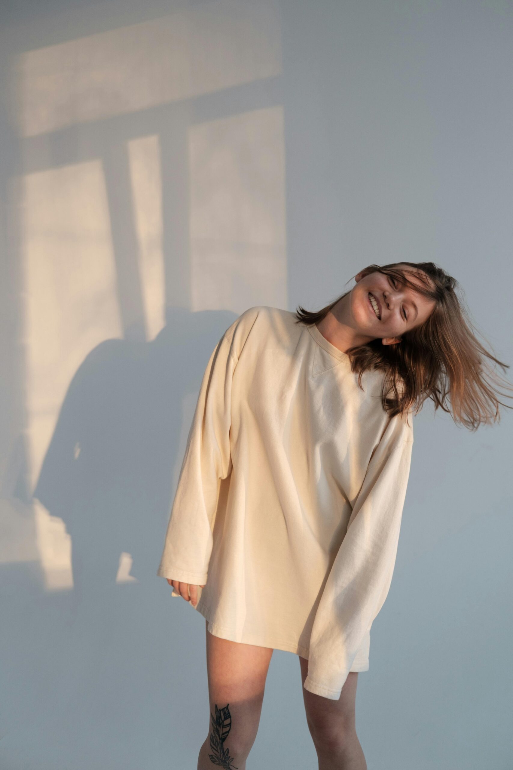 Happy young female in white sweatshirt smiling and shaking hair while dancing against gray wall
